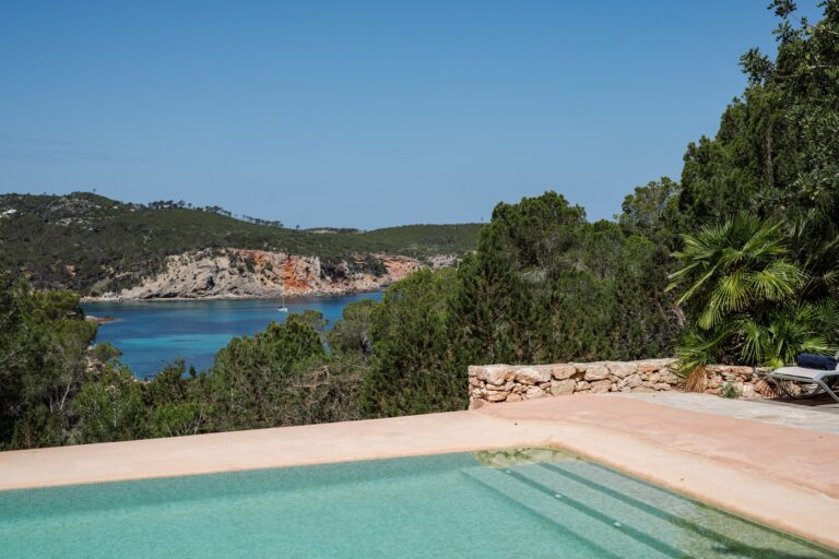 Pool edge overlooking forest, sea cove, rocky cliffs and palm trees at Casa Xarraca in San Juan, Ibiza