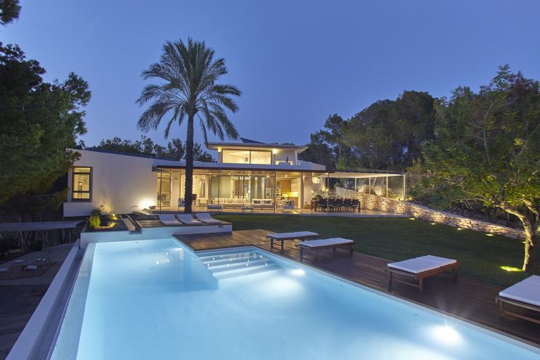 Modern villa at night with illuminated pool, palm tree and mature garden at Sol Vista, a luxury villa in Cala Salada, Ibiza