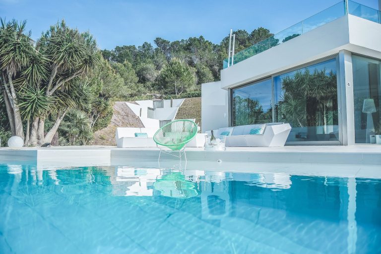 Pool terrace, white villa with glass railing, palm and pine trees at Villa Adelyn in San Agustin, Ibiza